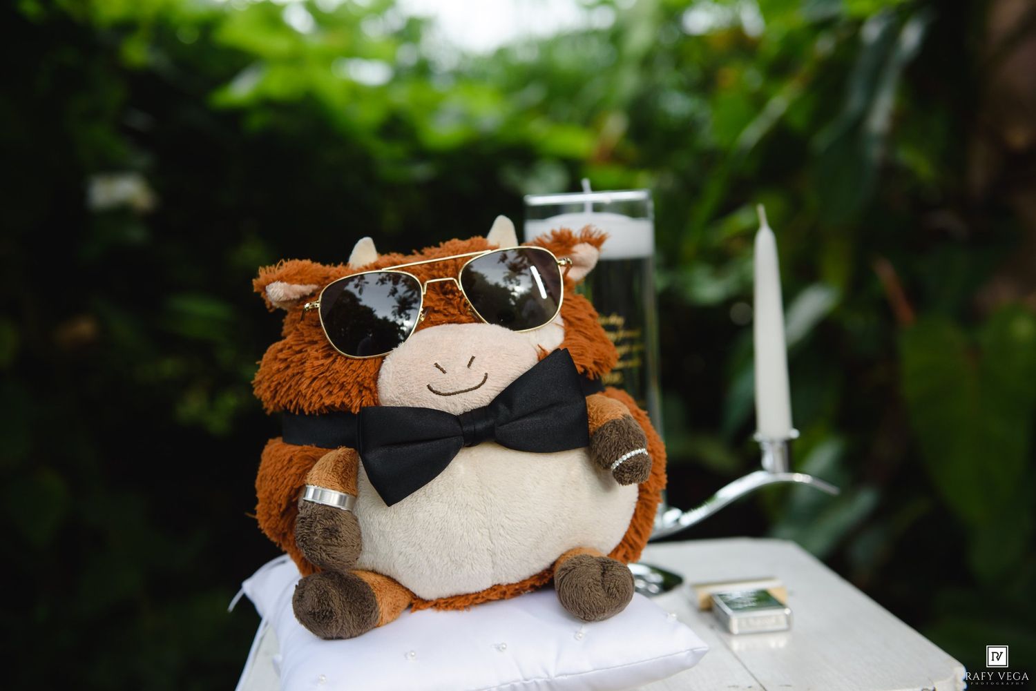 Cute stuffed cow toy wearing sunglasses and bow tie displayed on a white table outdoors.