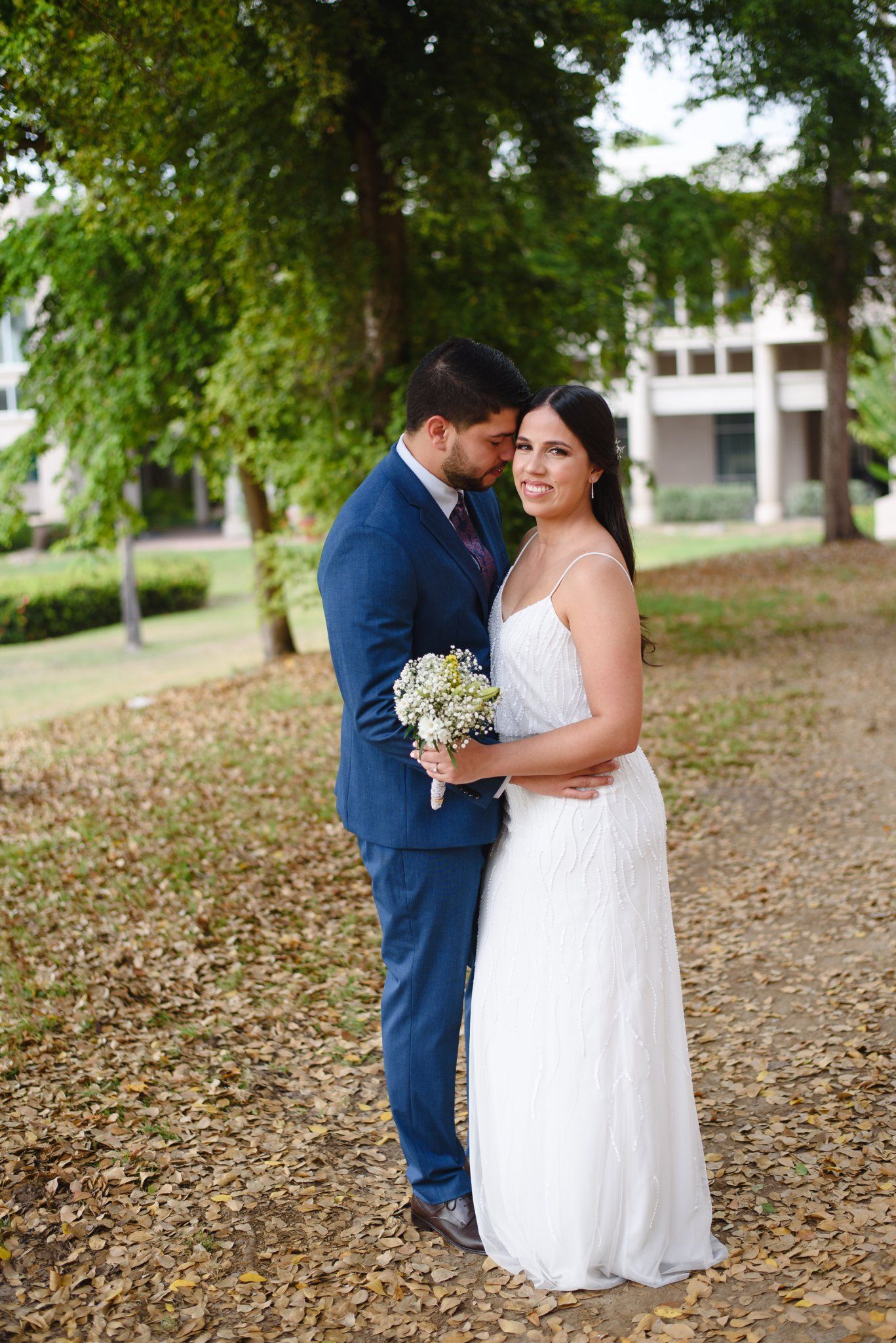 Bride in white dress holding bouquet embraces groom in blue suit outdoors among trees and fallen leaves.