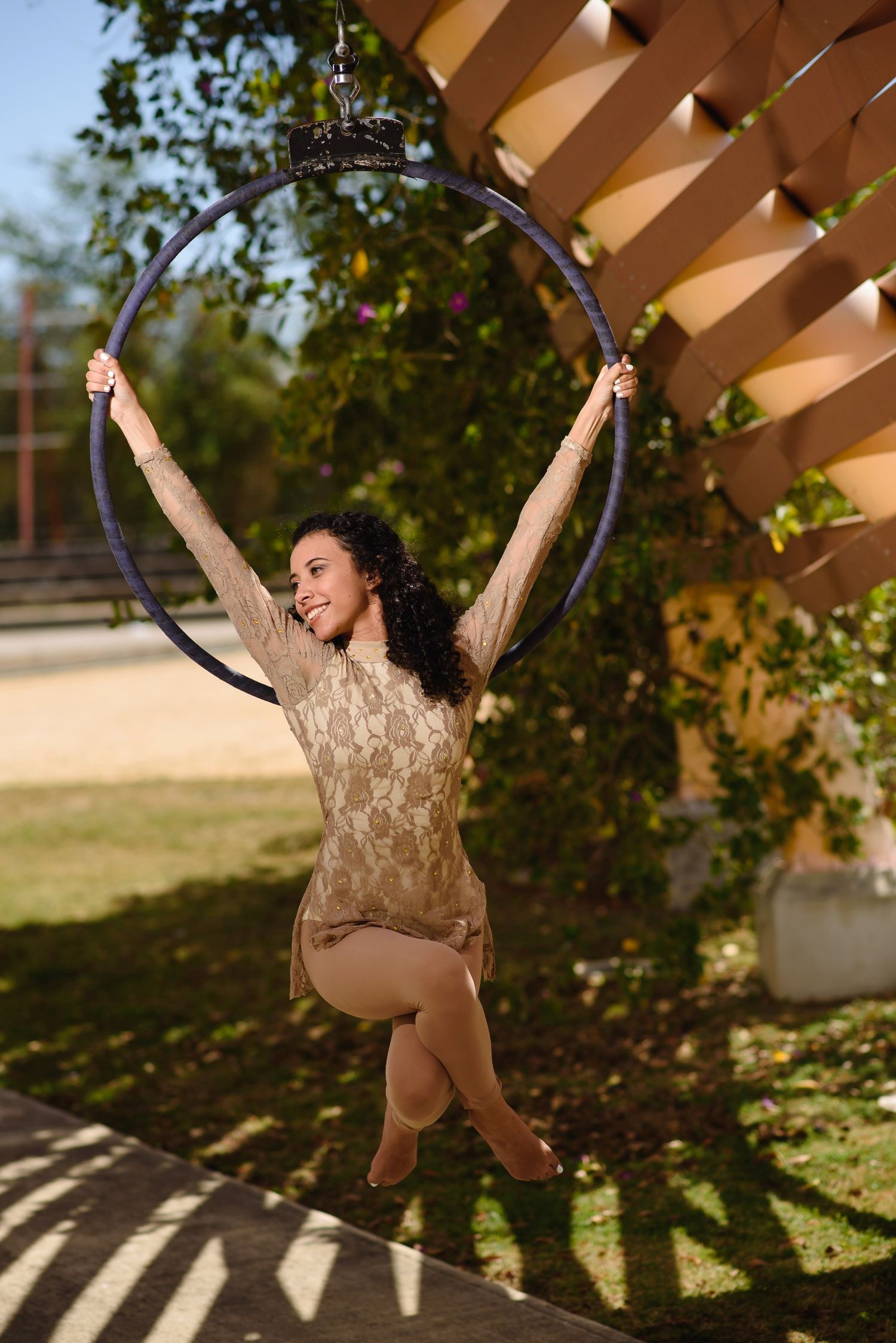 A smiling woman in a beige lace bodysuit performs aerial hoop acrobatics outdoors in a sunny garden setting.