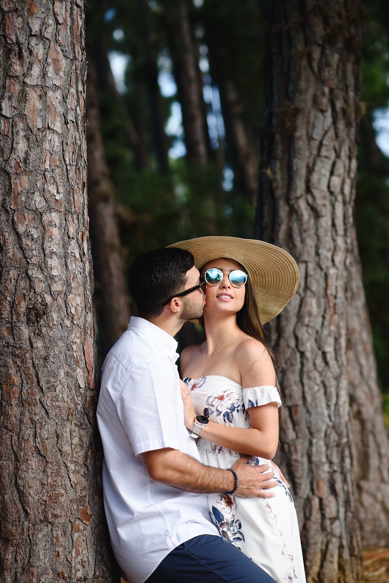 A couple embraces romantically among tall trees, woman wearing a wide-brim hat and sunglasses in a floral off-shoulder dress.