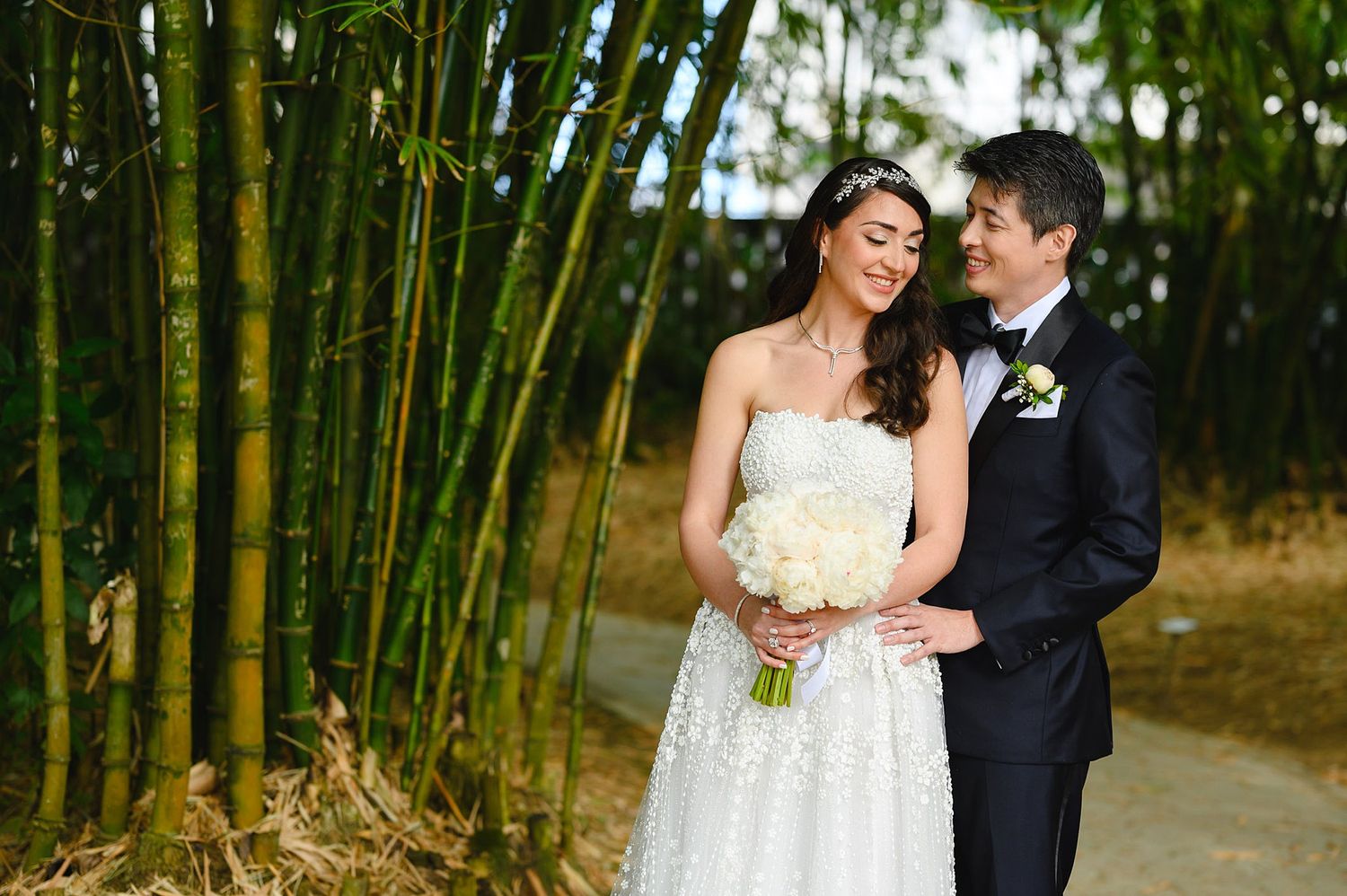 Happy bride and groom pose together in bamboo garden, bride in lace gown holding white floral bouquet.