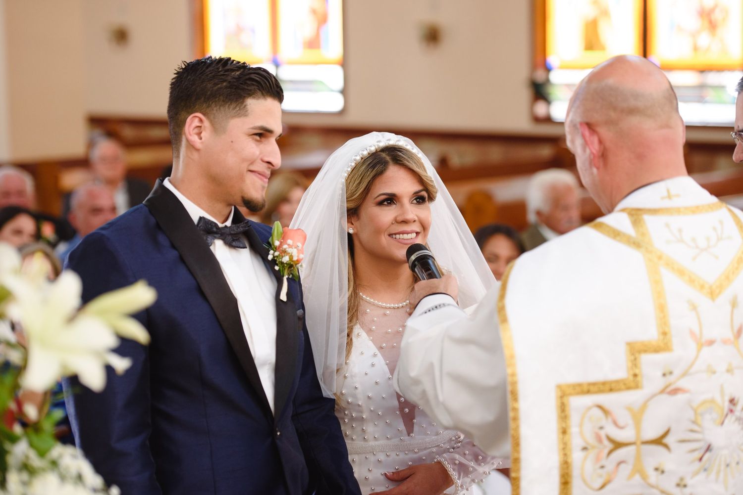Bride and groom exchange vows at church altar as priest holds microphone during wedding ceremony.