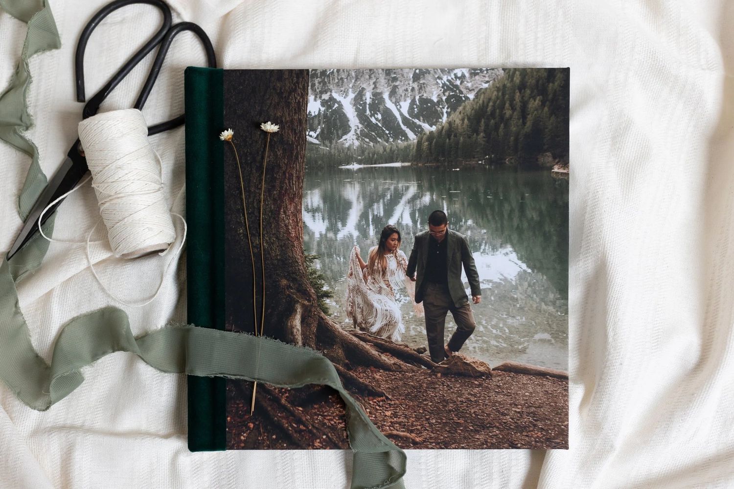 A wedding photo album with a forest cover image of a couple by a lake, surrounded by ribbon and twine.