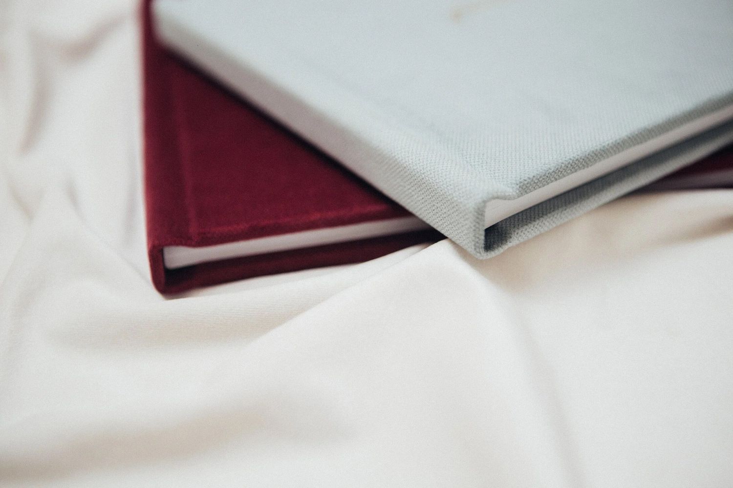 Two hardcover books in red and light blue lay stacked on white fabric.