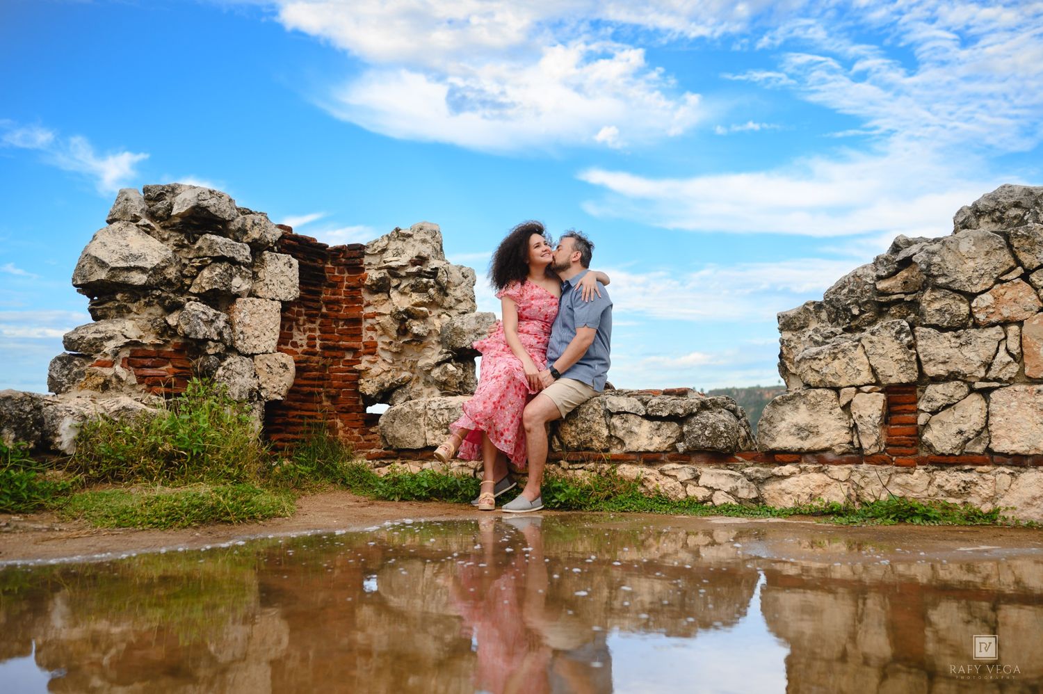 Romantic couple kissing among ancient stone ruins with blue sky and puddle reflection.