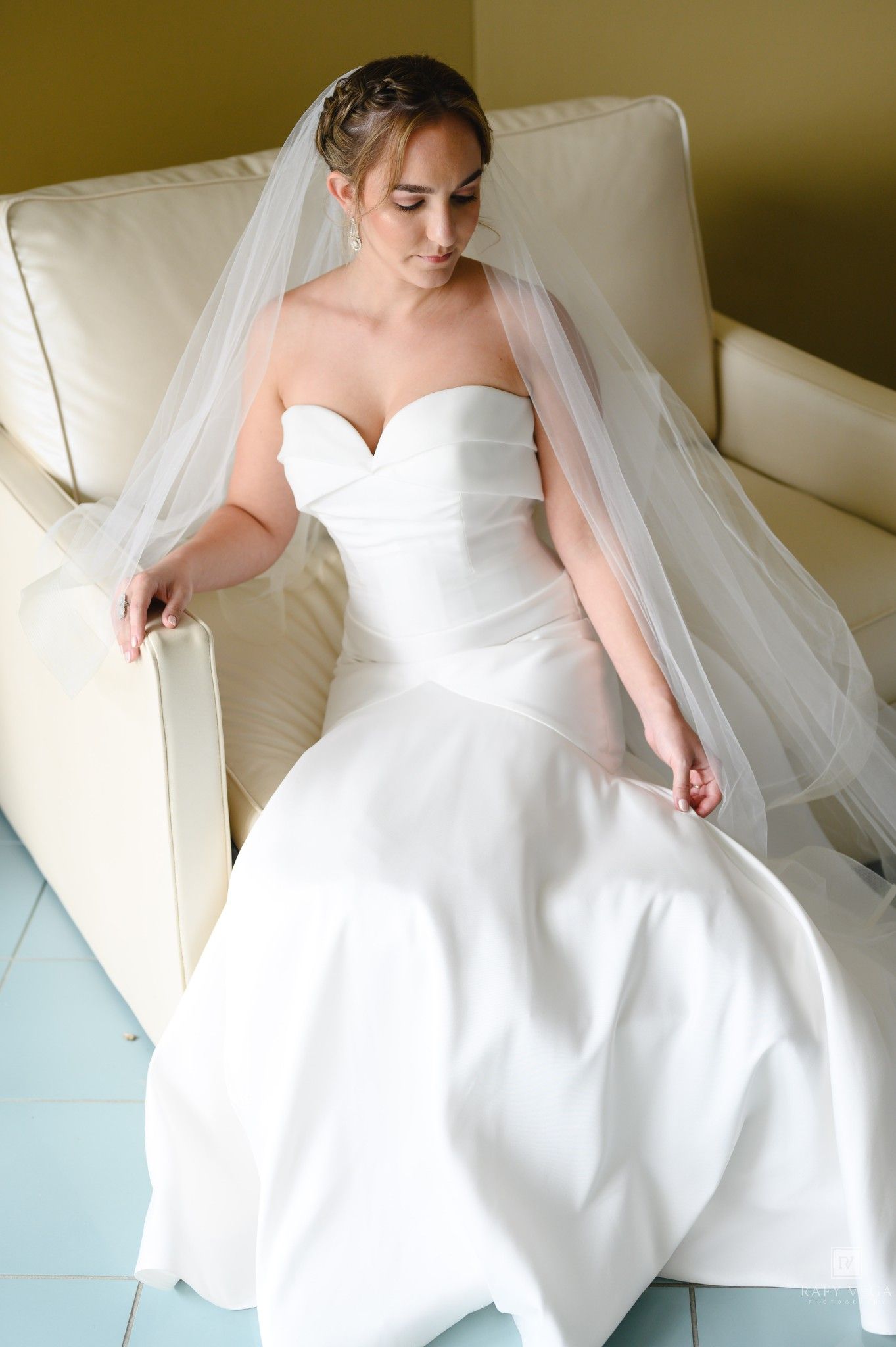Bride in strapless white wedding gown and veil seated gracefully on cream leather chair.