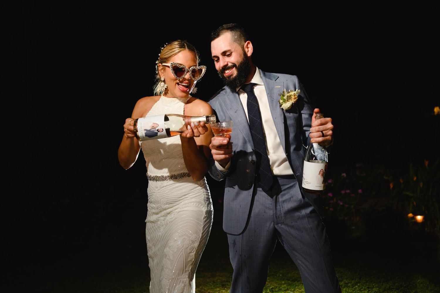 Bride and groom laughing together at night, holding drinks and wearing fun sunglasses at their wedding reception.