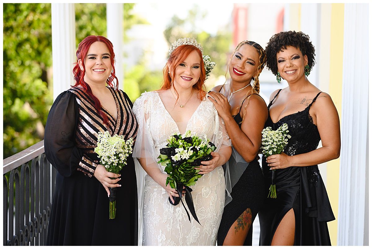Bride in white gown with three bridesmaids in black dresses holding bouquets on a decorated balcony.