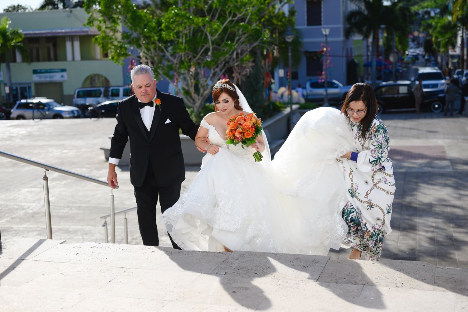 Bride in white gown with orange bouquet escorted up steps by man in tuxedo and woman at wedding.