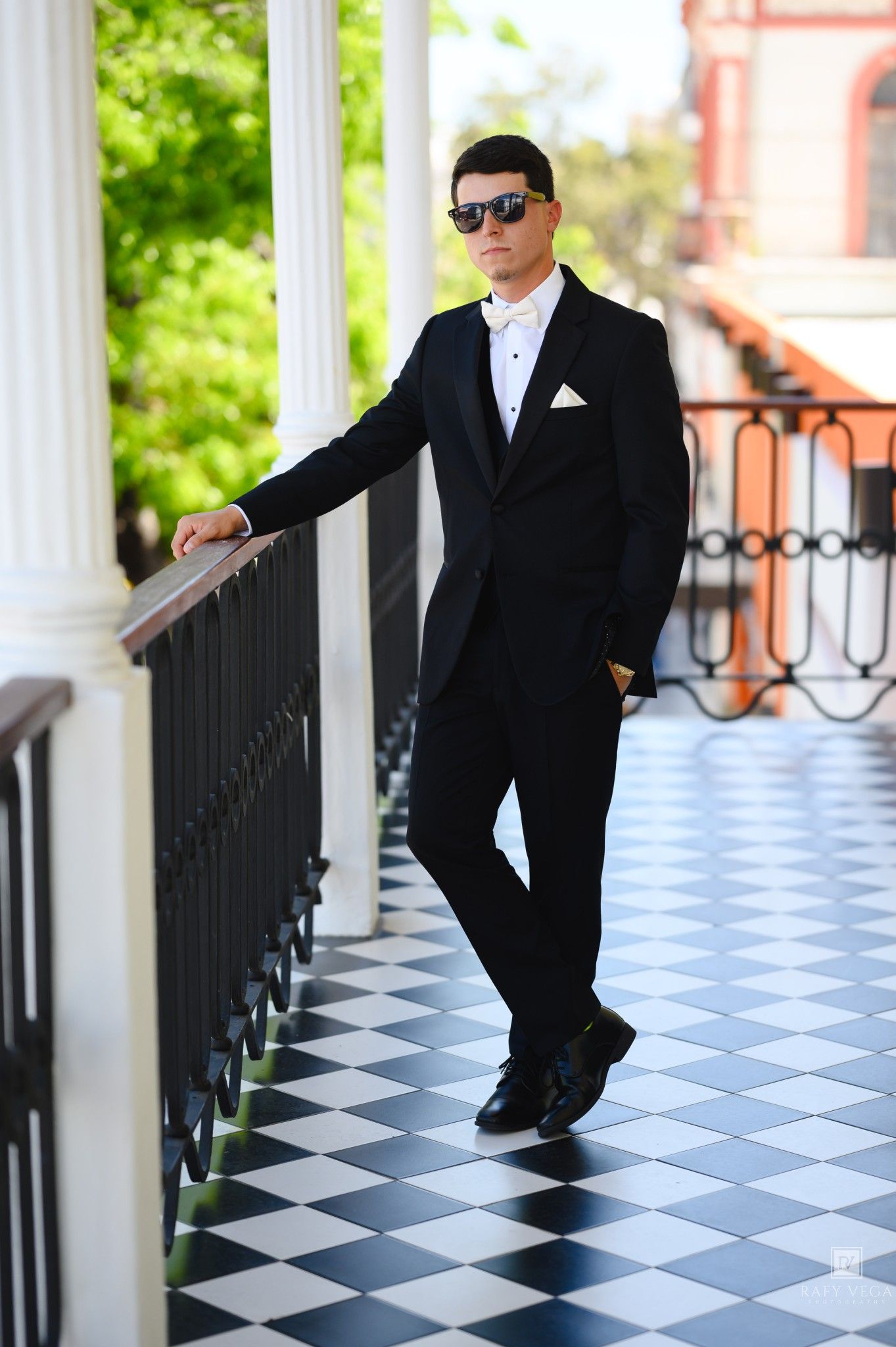 Young man in black tuxedo with white bow tie and sunglasses posing on checkered floor balcony.