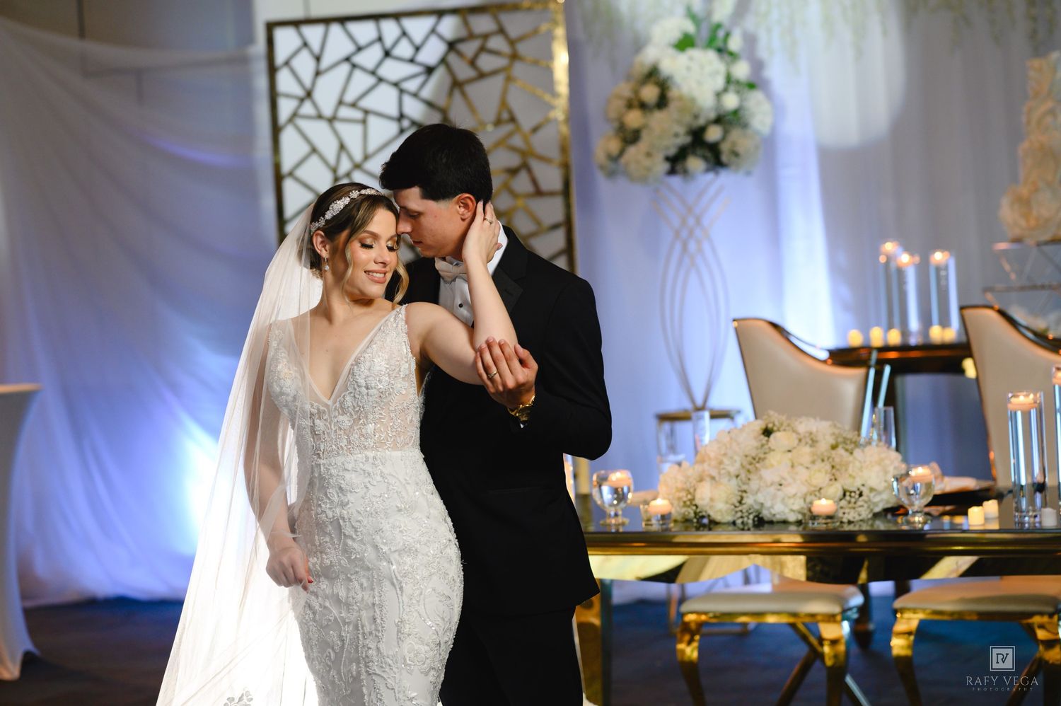 Bride and groom share a romantic moment at elegant wedding reception with white floral decor and draped backdrop.