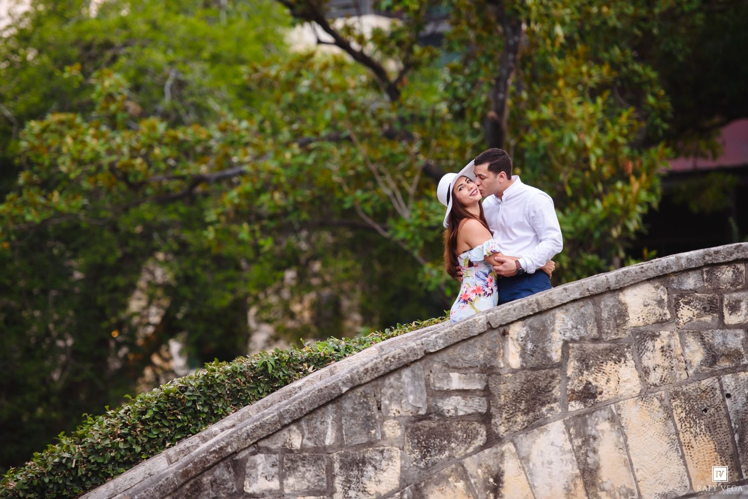 Romantic couple sharing a kiss on a stone bridge surrounded by lush green trees in an outdoor setting.