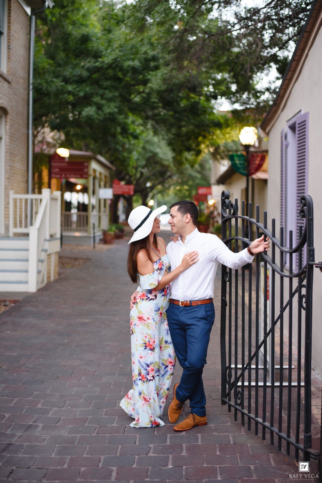 Couple in floral dress and white hat embracing by iron gate on charming brick street.