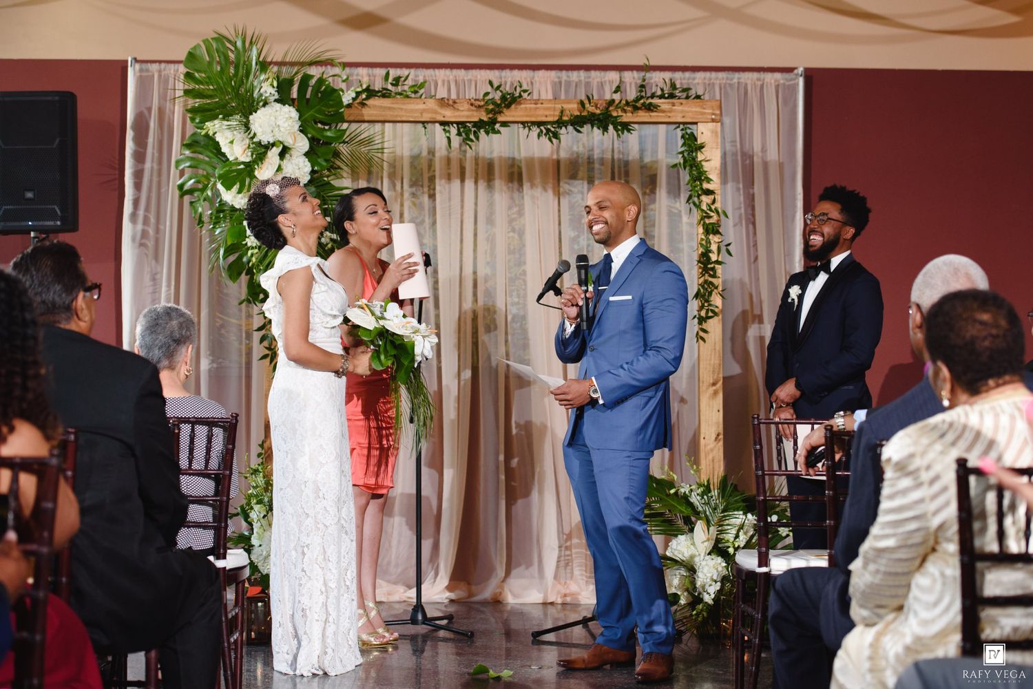 Couple exchanging vows at elegant wedding ceremony beneath floral arch with guests seated in background.