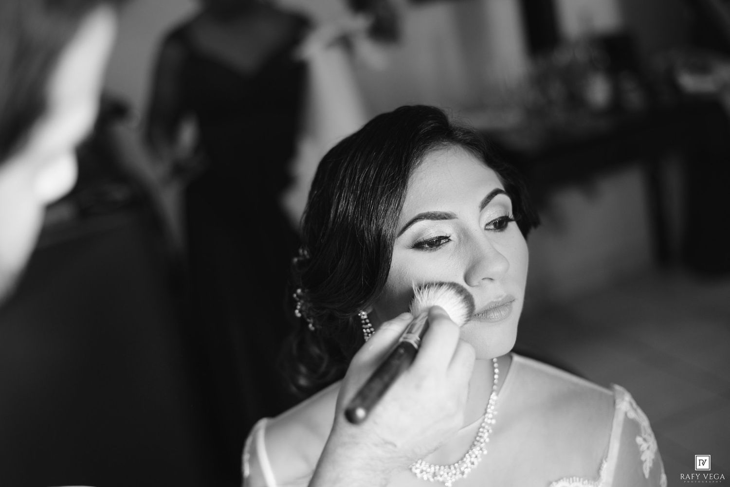Black and white photo of a bride getting makeup applied with a brush on her cheek before her wedding ceremony.