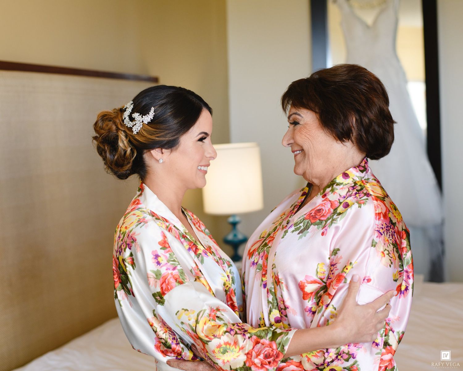 Bride and mother sharing a joyful embrace in matching floral satin robes during wedding morning preparations.