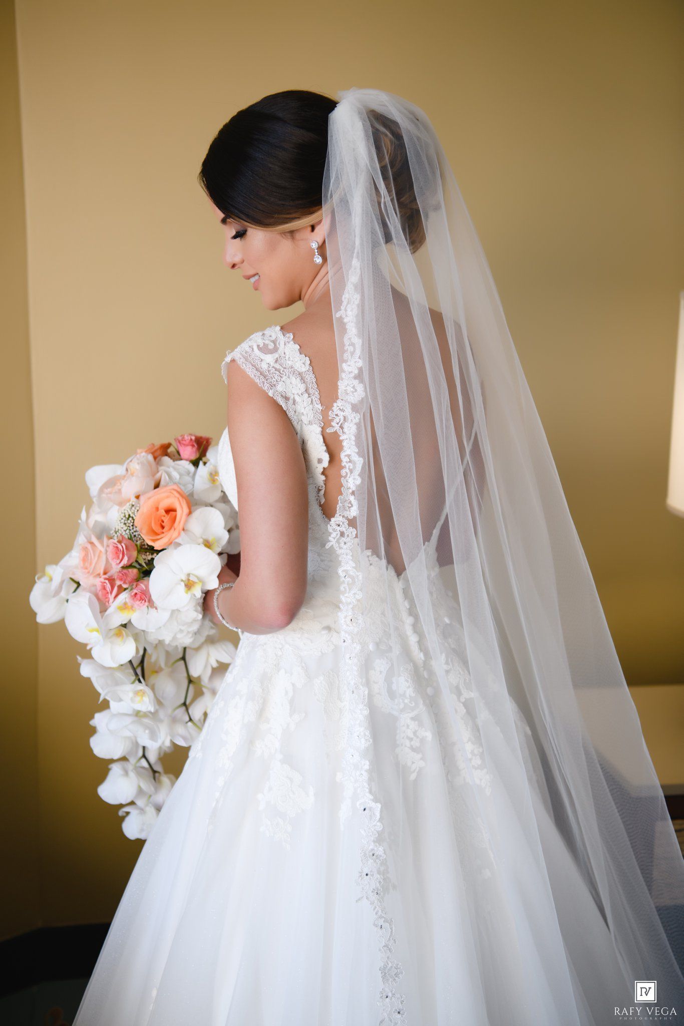 Bride in backless lace wedding gown with long veil holding a cascading pink and white floral bouquet.