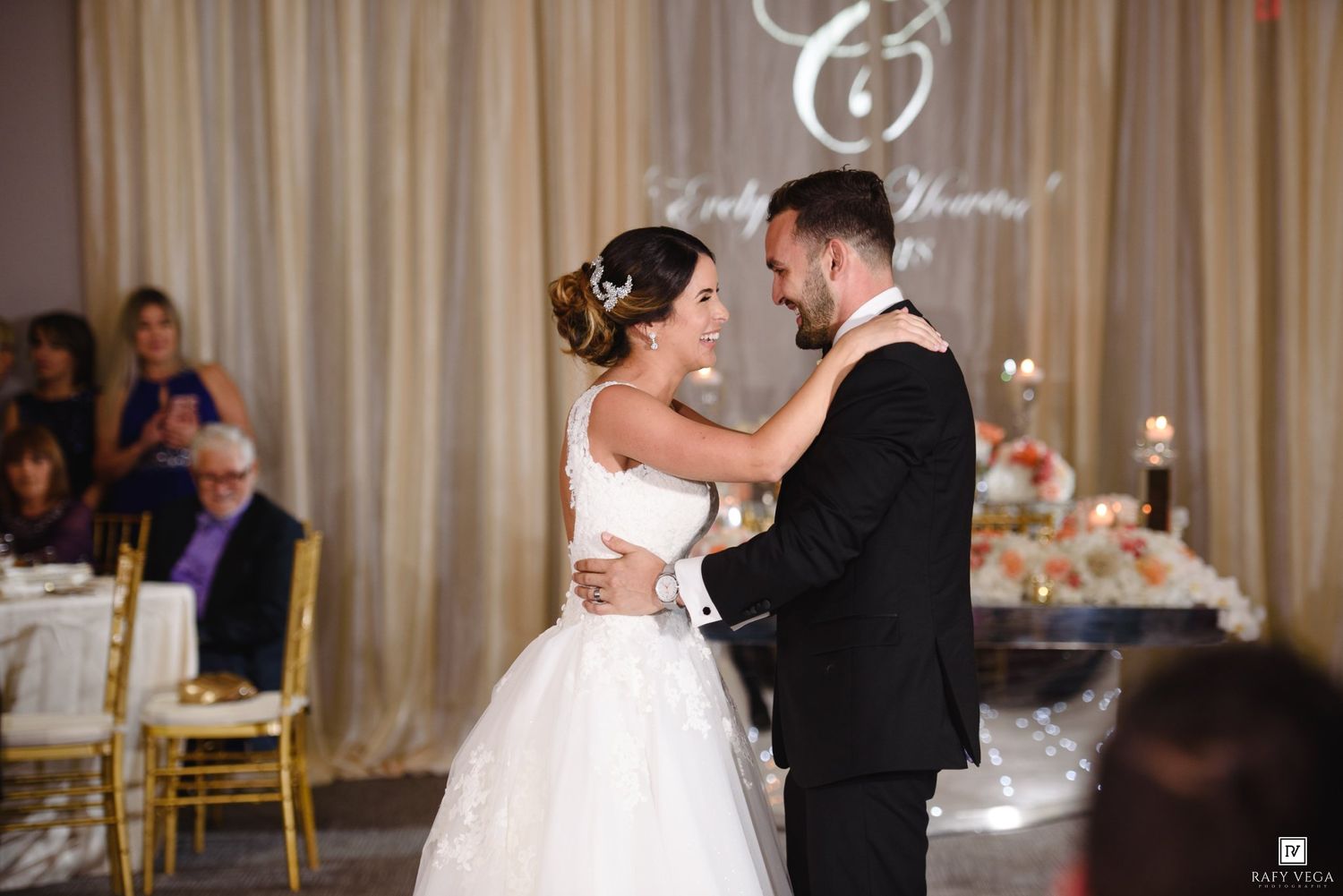 Bride and groom share their first dance at elegant wedding reception with floral decorations and guests in background.