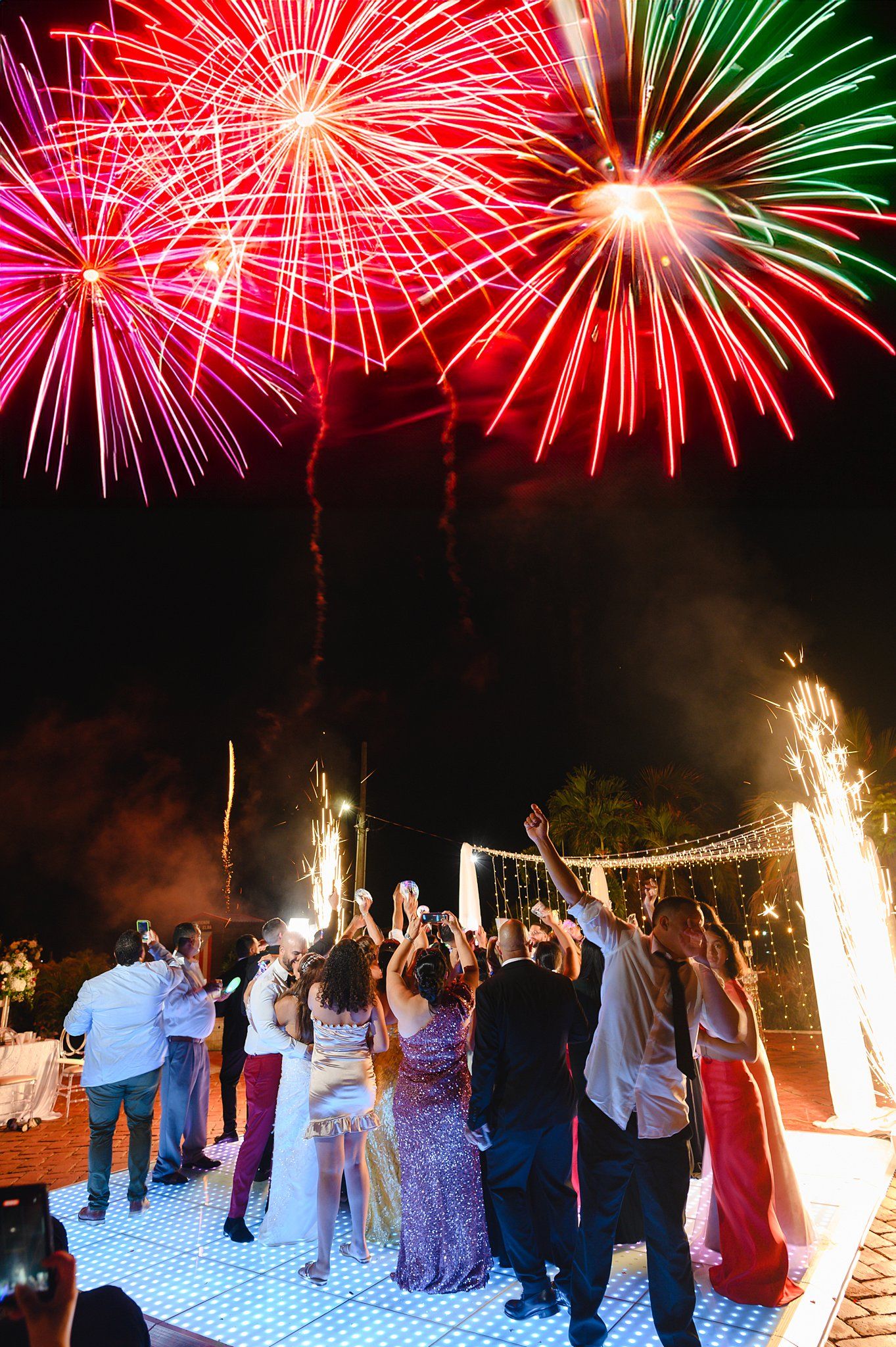 Crowd of guests celebrating outdoors at night with vibrant red and green fireworks exploding in the sky above.