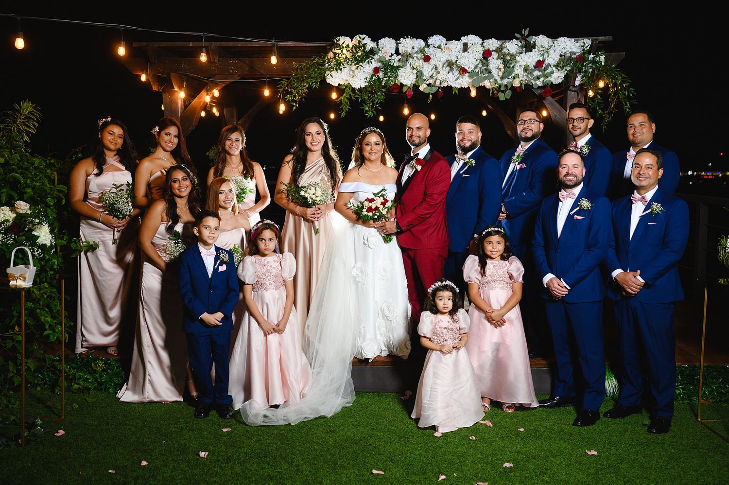 Wedding party poses outdoors at night under floral arch with string lights, bride in white, groom in red, groomsmen in blue.