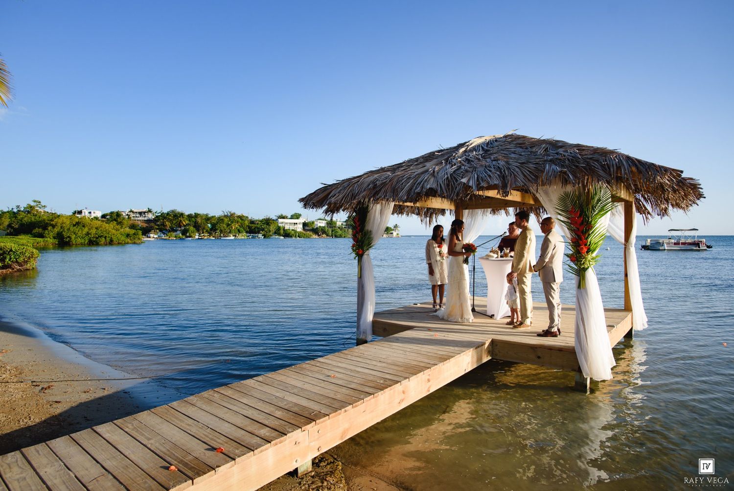 Tropical overwater gazebo on a wooden dock with a thatched roof, surrounded by calm blue waters under a clear sky.
