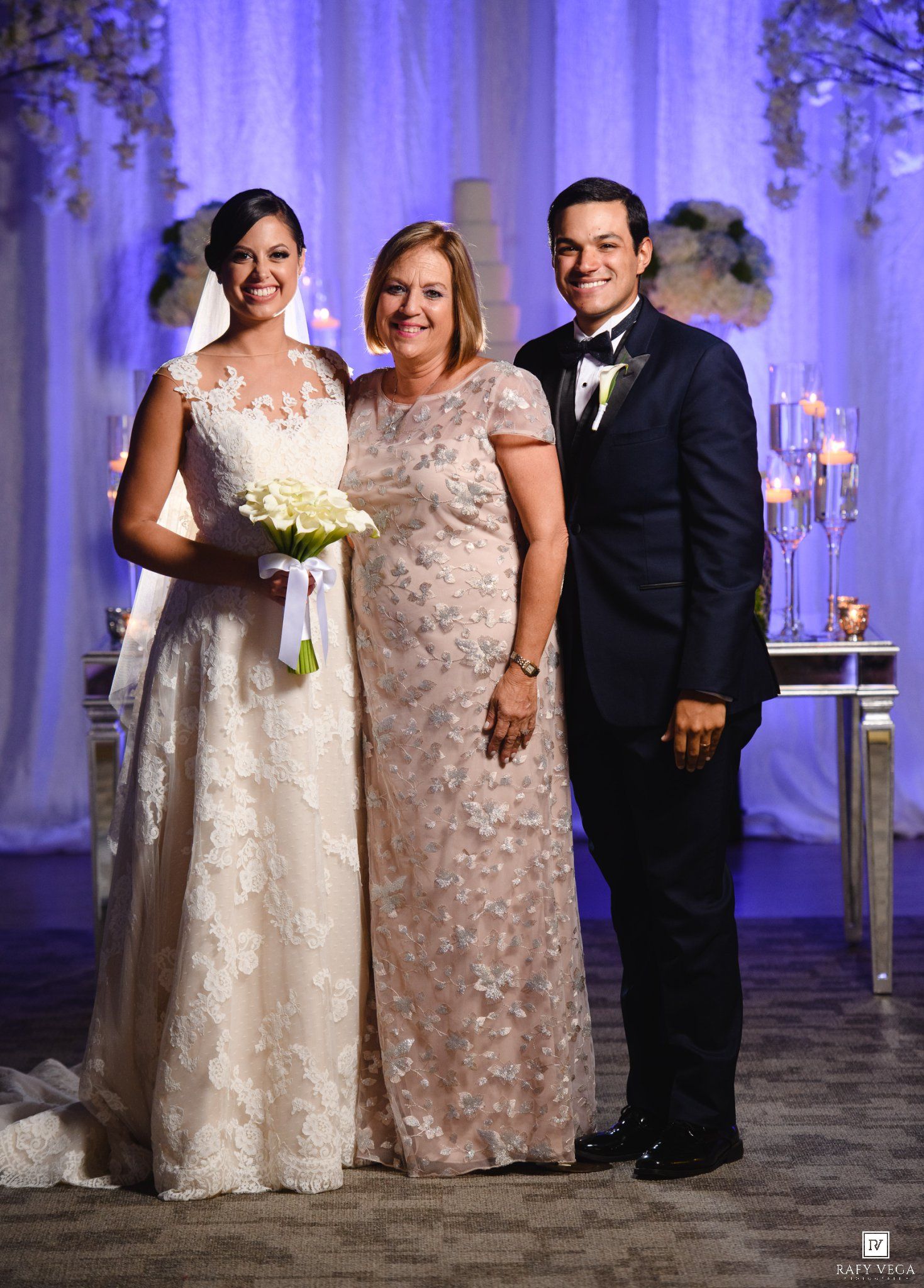 Bride in white lace gown and groom in navy tuxedo pose with older woman in floral dress at elegant wedding venue.