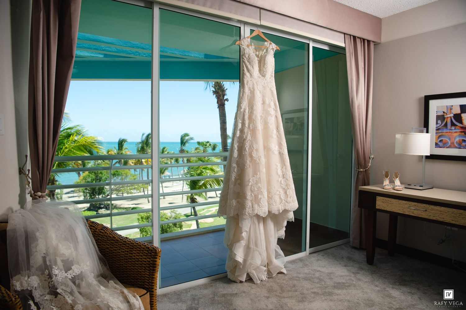 White lace wedding dress hanging on glass door with ocean view balcony and palm trees in tropical hotel room.