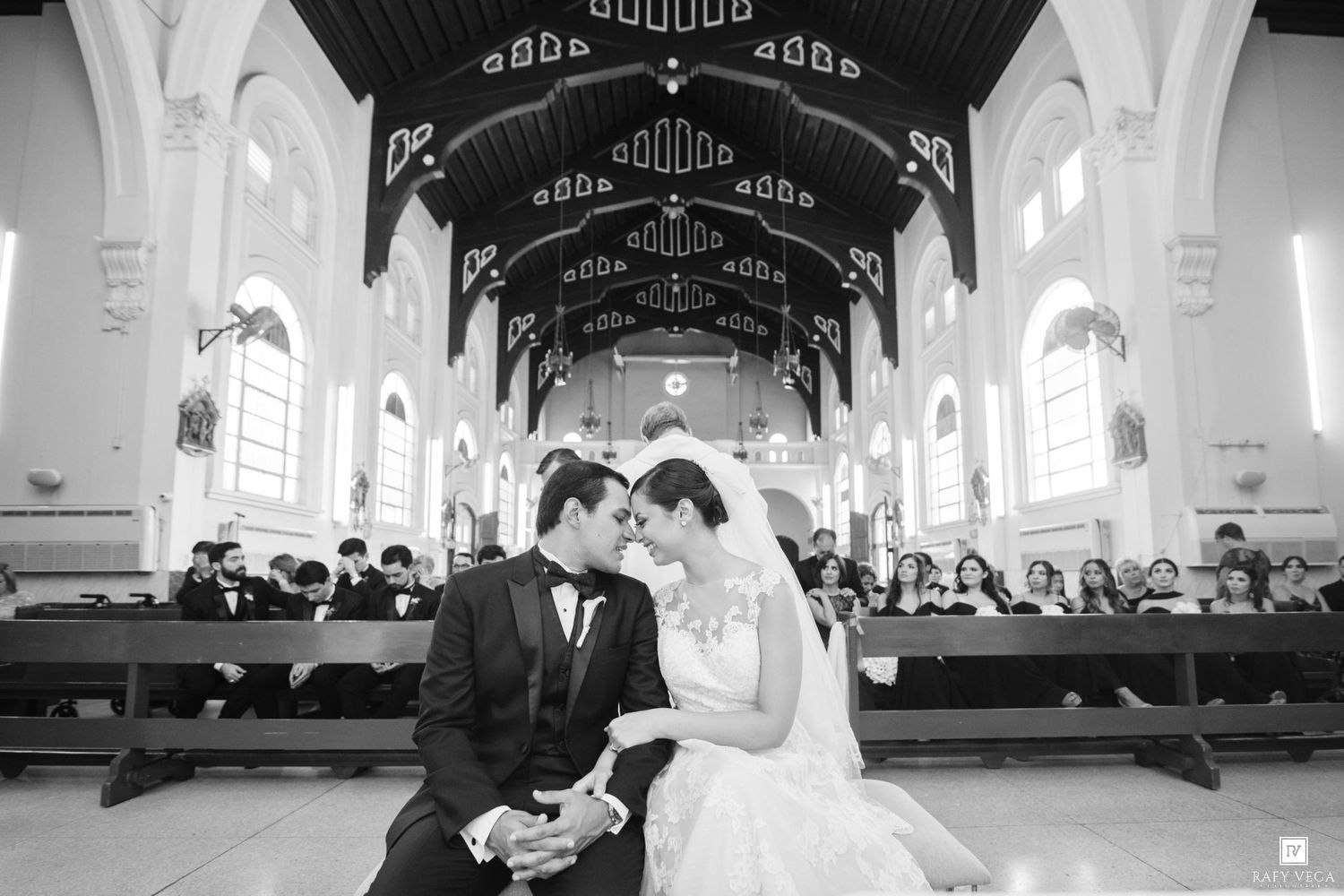 Black and white wedding photo of bride and groom sharing intimate moment in elegant church aisle.