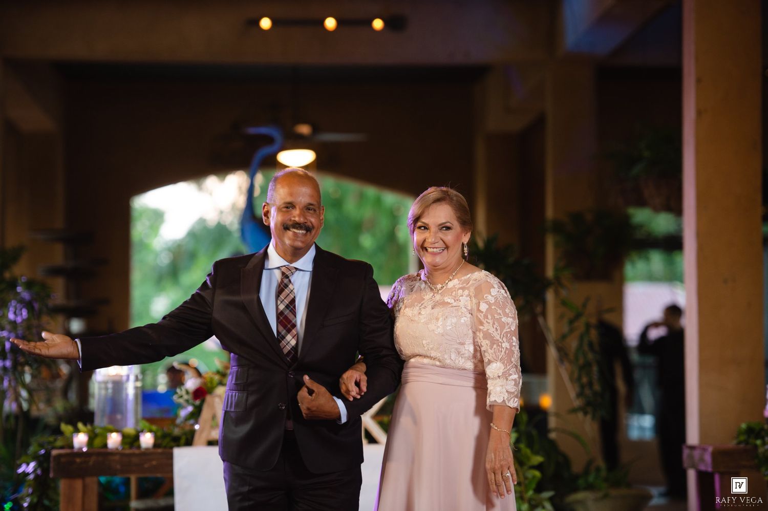 Elegant couple smiling at a wedding reception, man in suit and woman in lace dress, standing in decorated venue.