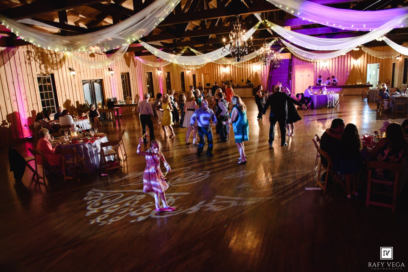 Guests dancing on a wooden dance floor at an elegant wedding reception with pink uplighting and draped ceiling decor.