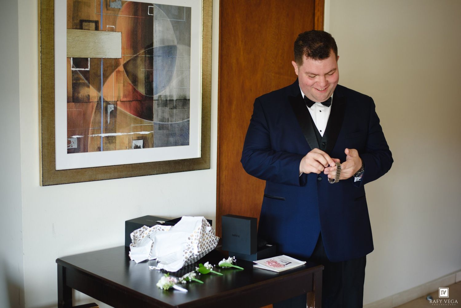 Groom in navy blue suit adjusting bow tie before wedding ceremony in elegant hotel room.