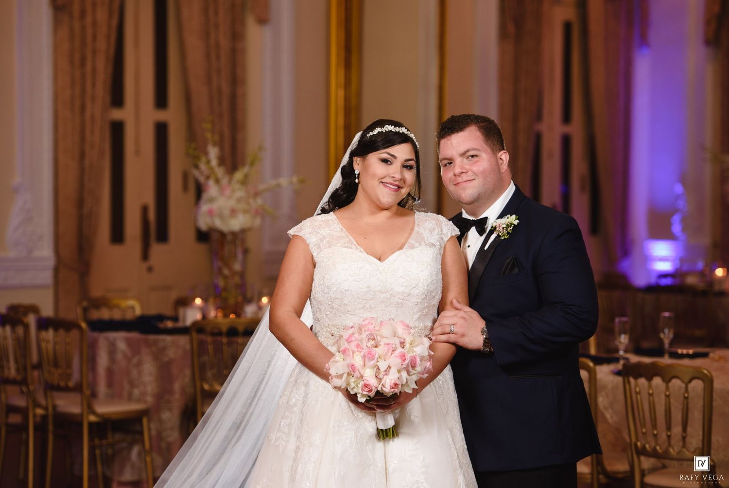 Bride in white ball gown holding pink bouquet stands with groom in navy tuxedo at elegant wedding reception.