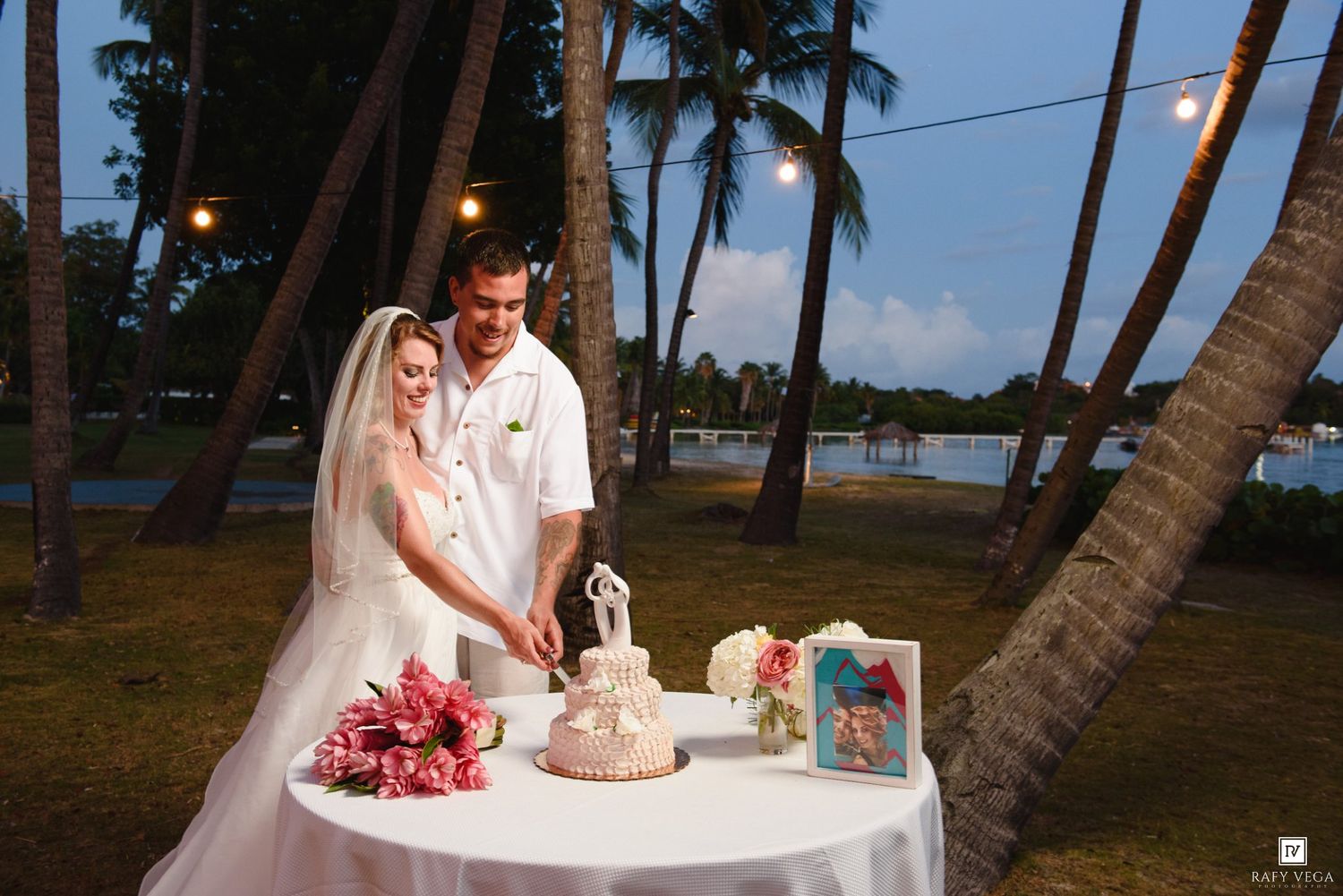 Bride and groom cut their wedding cake outdoors under palm trees with string lights at dusk.