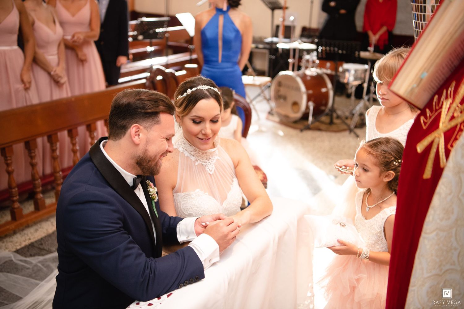 Bride and groom exchanging rings at church wedding ceremony with flower girl and guests in background.
