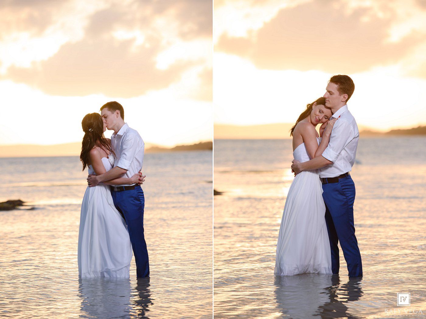 Couple kissing and embracing at sunset on a beach, bride in white gown and groom in blue pants standing in shallow water.