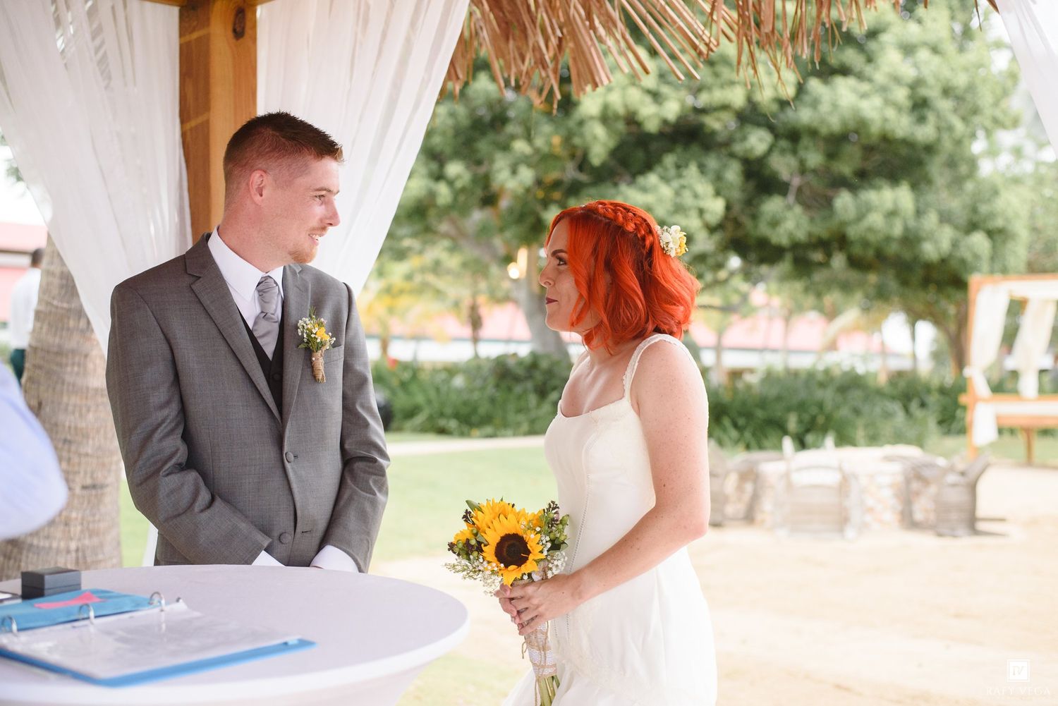 Bride holding sunflowers and groom in gray suit exchange vows at an outdoor wedding ceremony under a thatched canopy.