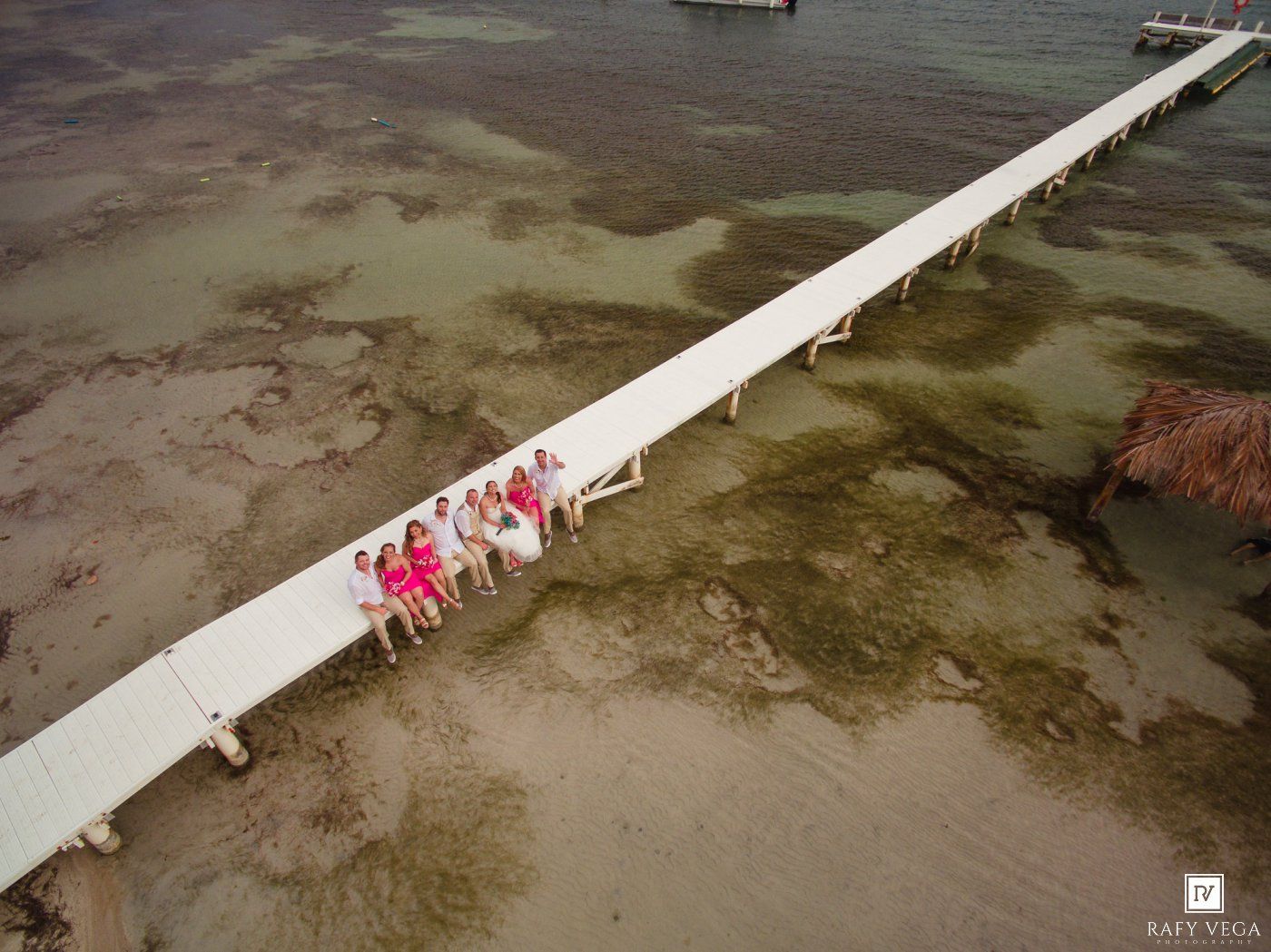Aerial view of a wedding party in colorful dresses gathered on a long white pier extending over shallow coastal waters.