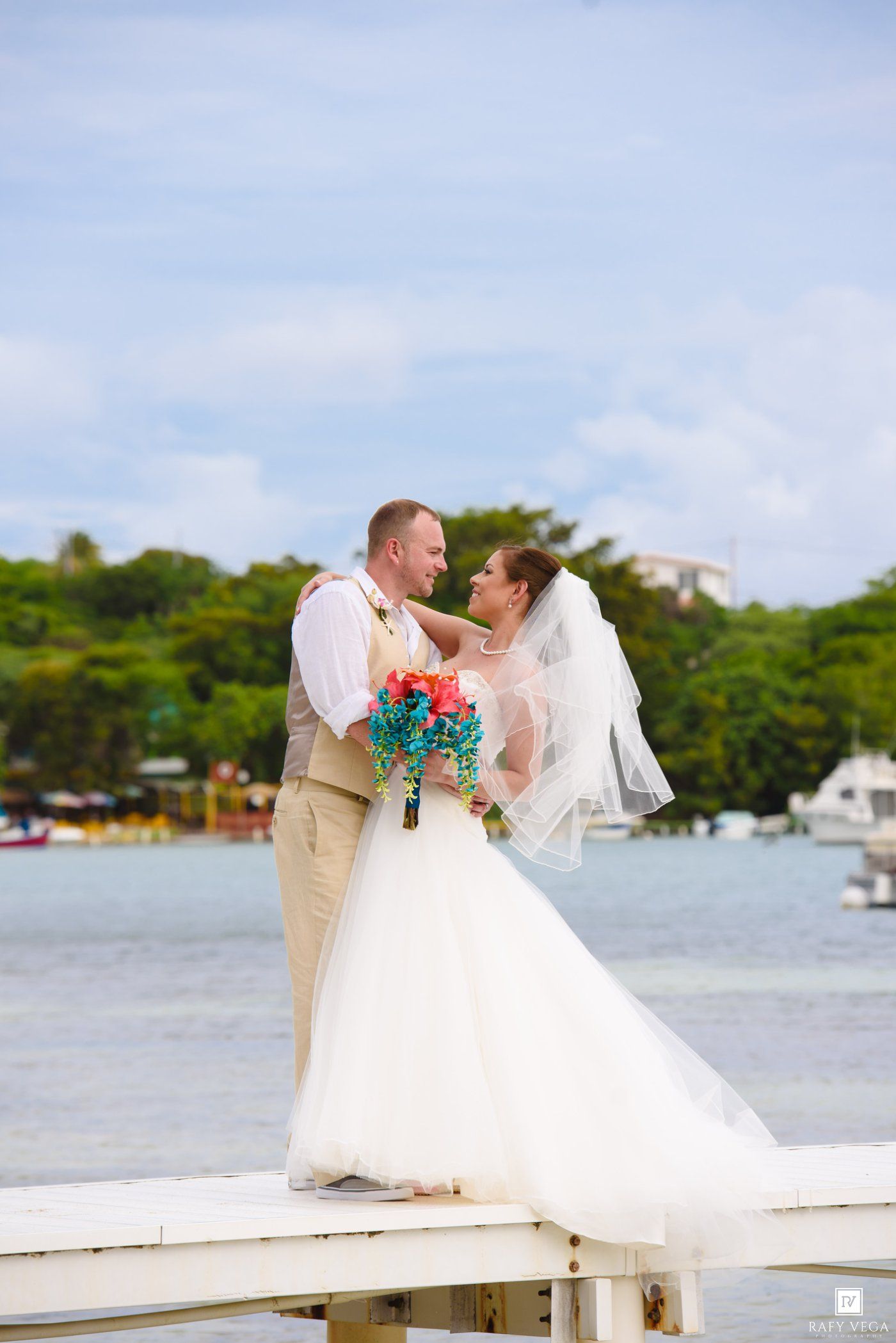Bride and groom share a romantic moment on a white dock overlooking a tropical bay, bride holding colorful bouquet.