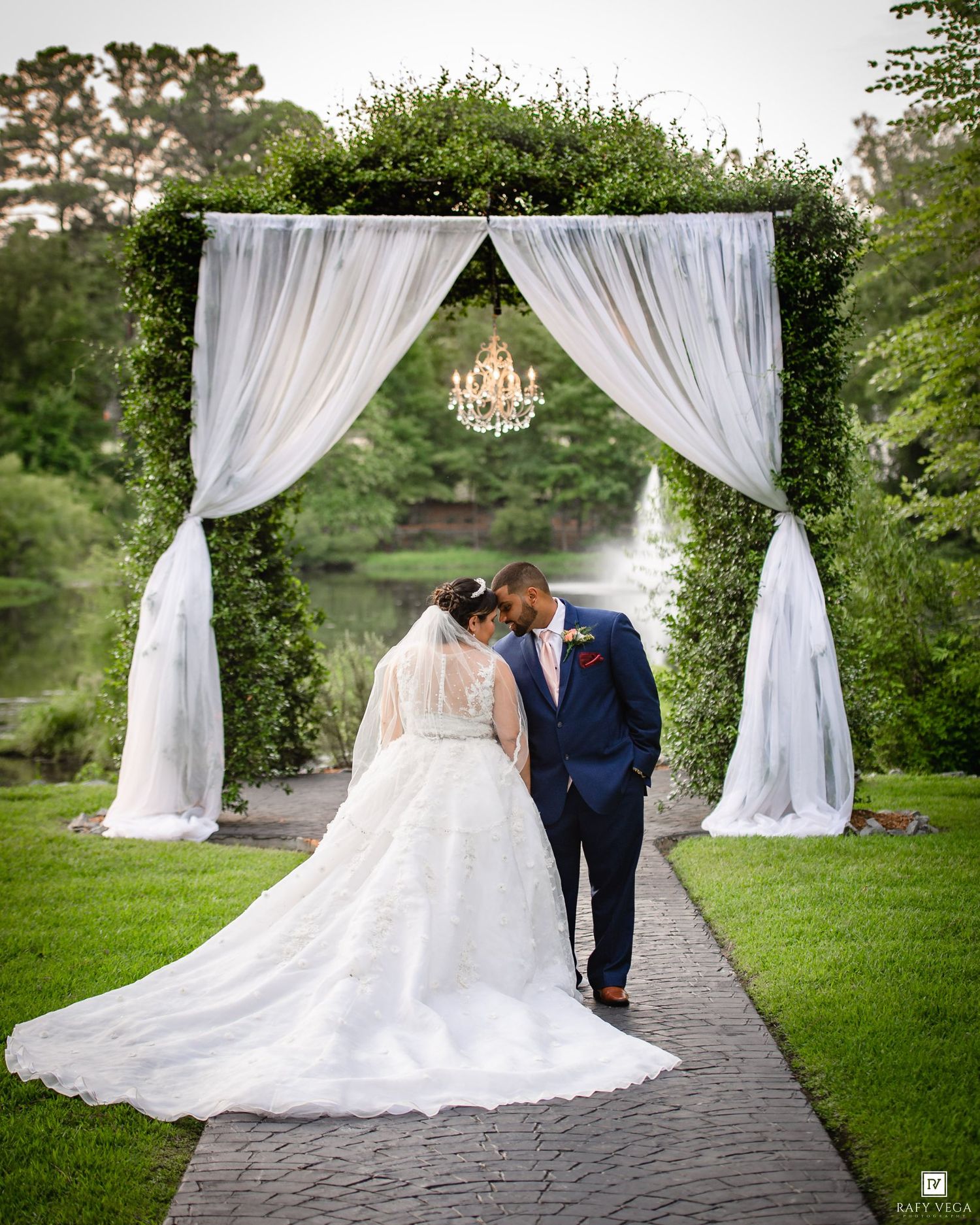 Bride and groom share a romantic moment under a floral arch with white drapes and chandelier at an outdoor wedding.