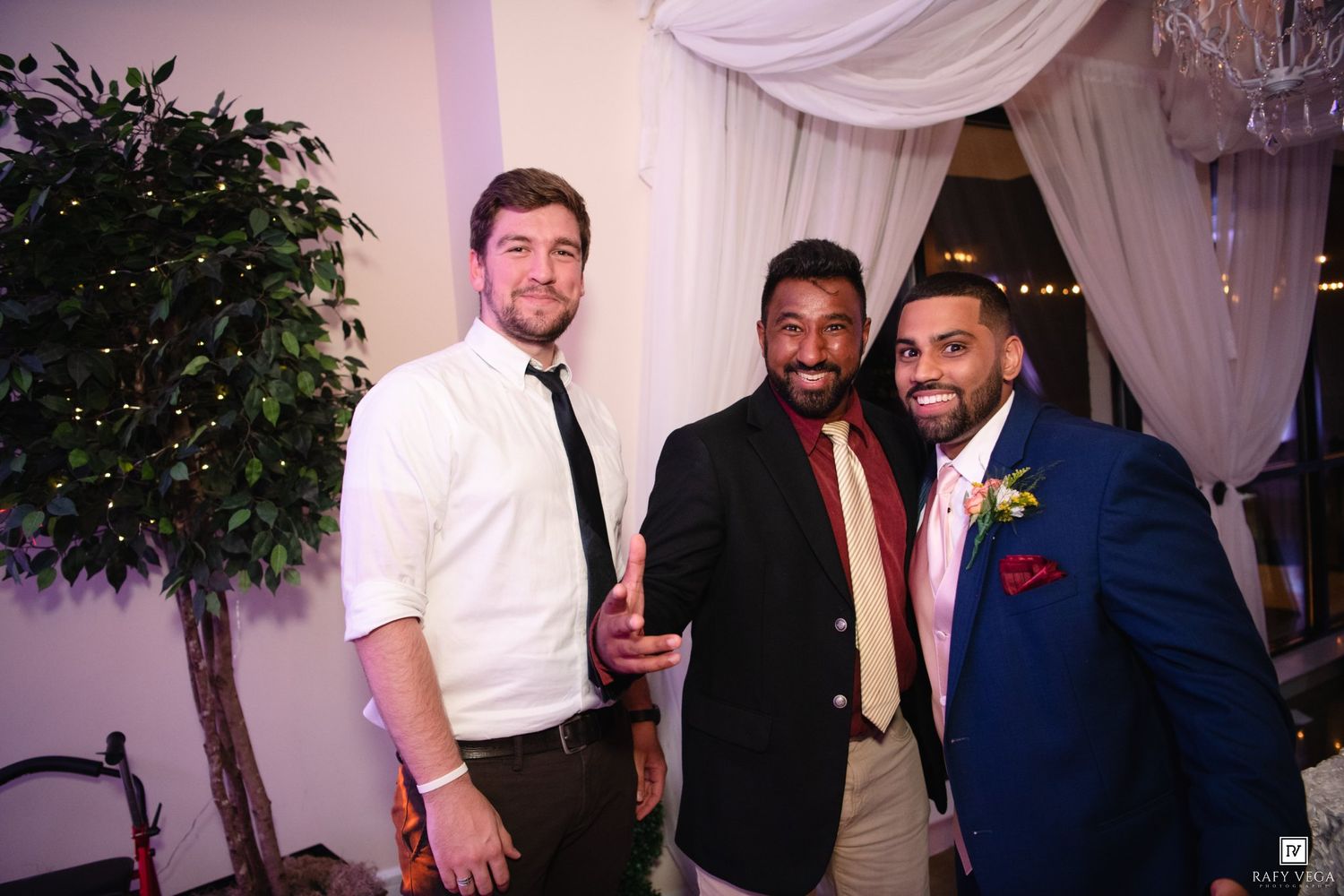 Three men in formal attire smile together at an elegant wedding reception venue with draped curtains.