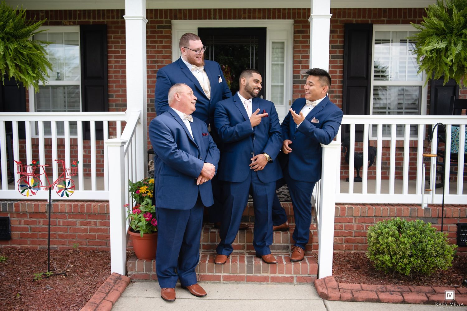 Four men in navy blue suits gathered on brick porch steps of a house, laughing and conversing at a wedding.