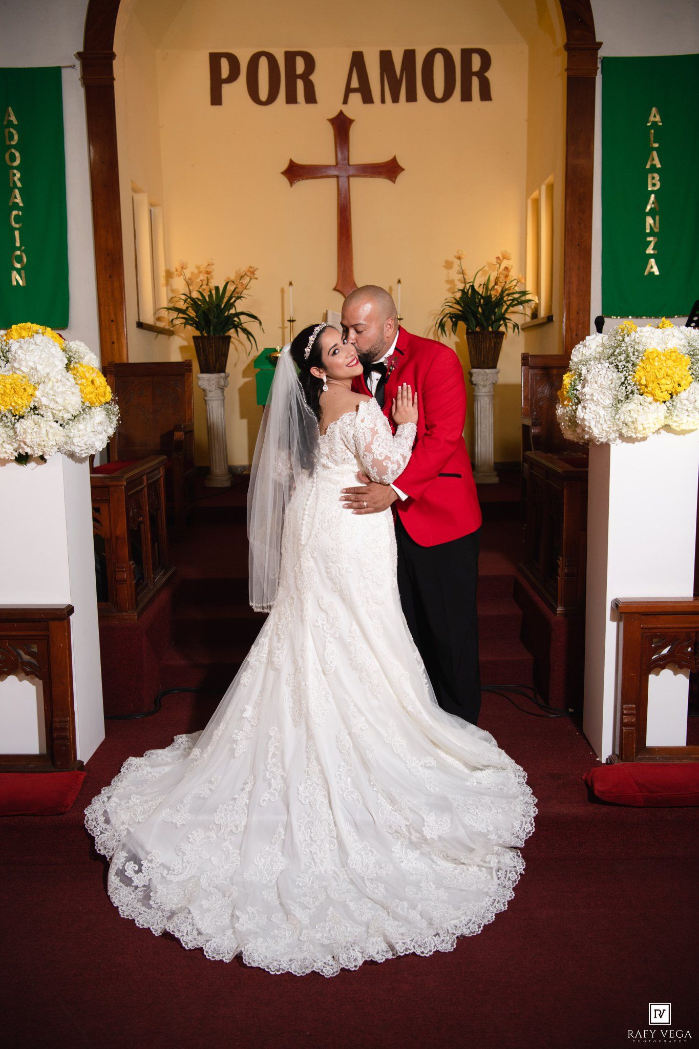 Bride in lace gown and groom in red tuxedo share a romantic kiss at church altar with cross and floral arrangements.
