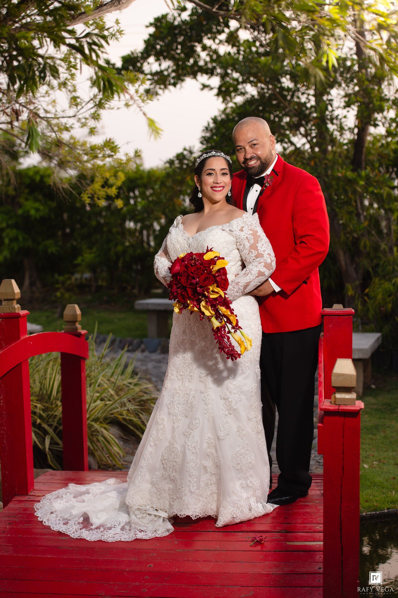 Bride and groom pose on red bridge, she holds red bouquet in lace gown, he wears red tuxedo jacket.