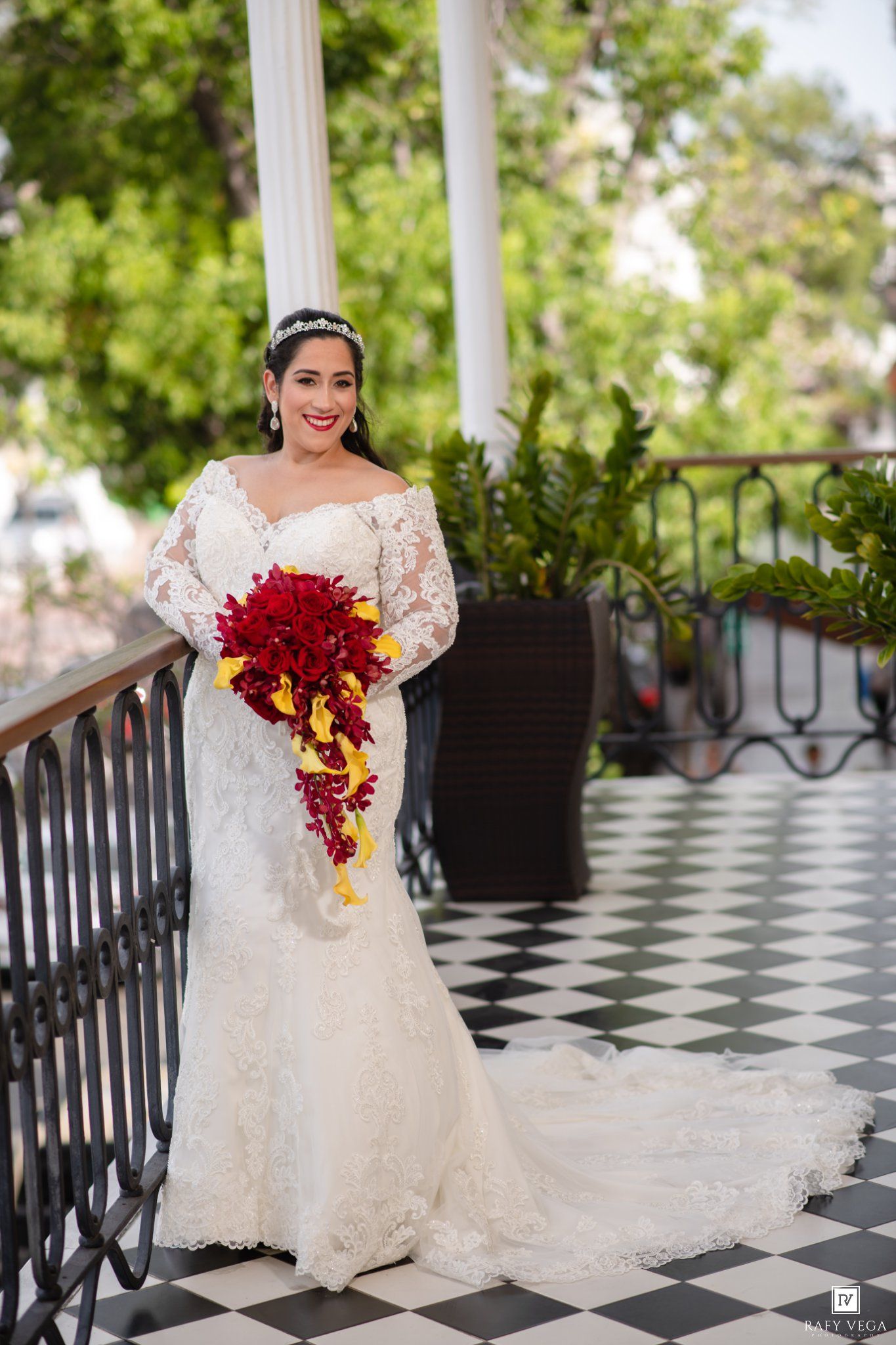 Bride in white lace mermaid gown holding red cascading bouquet on checkered balcony with greenery backdrop.