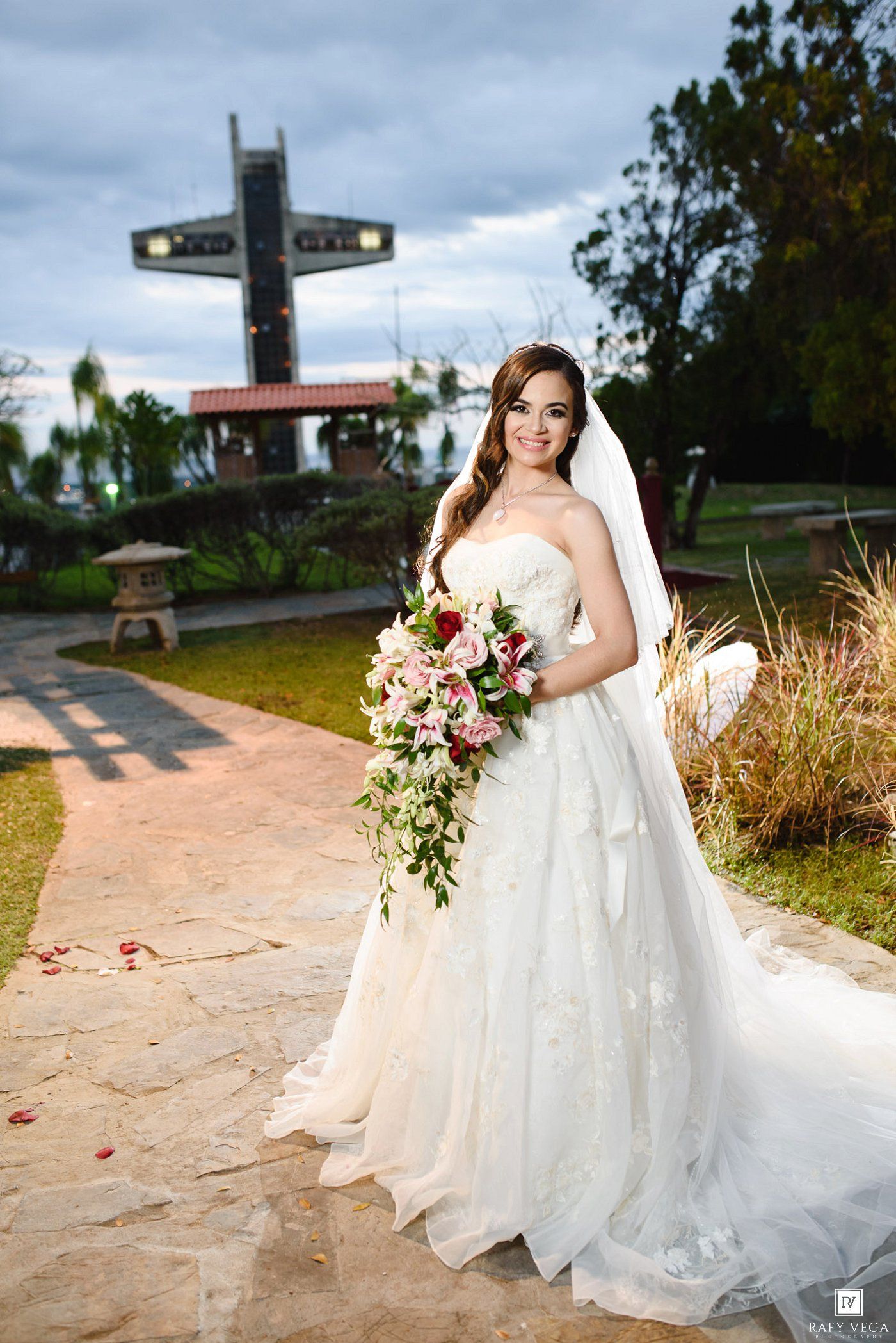 Bride in white strapless gown holding cascading floral bouquet stands on stone path with cross monument backdrop.
