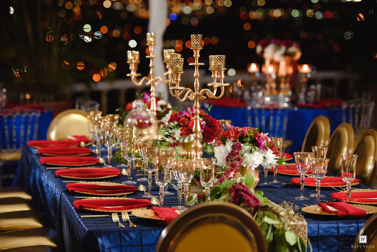 Elegant wedding reception table with blue linens, gold candelabras, red napkins, crystal glasses, and floral centerpieces at night.