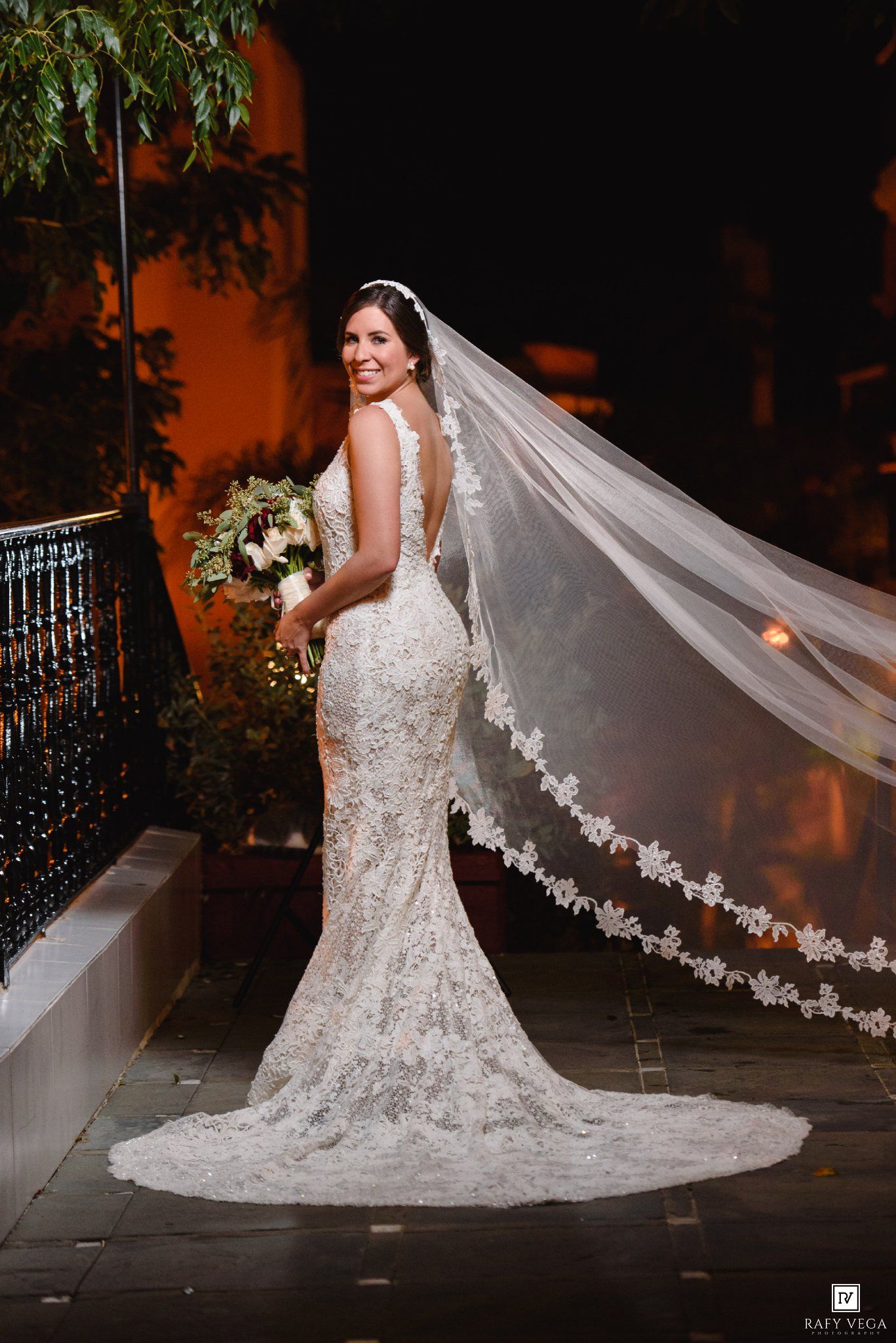Bride in lace mermaid wedding gown with cathedral veil and bouquet poses outdoors at night.