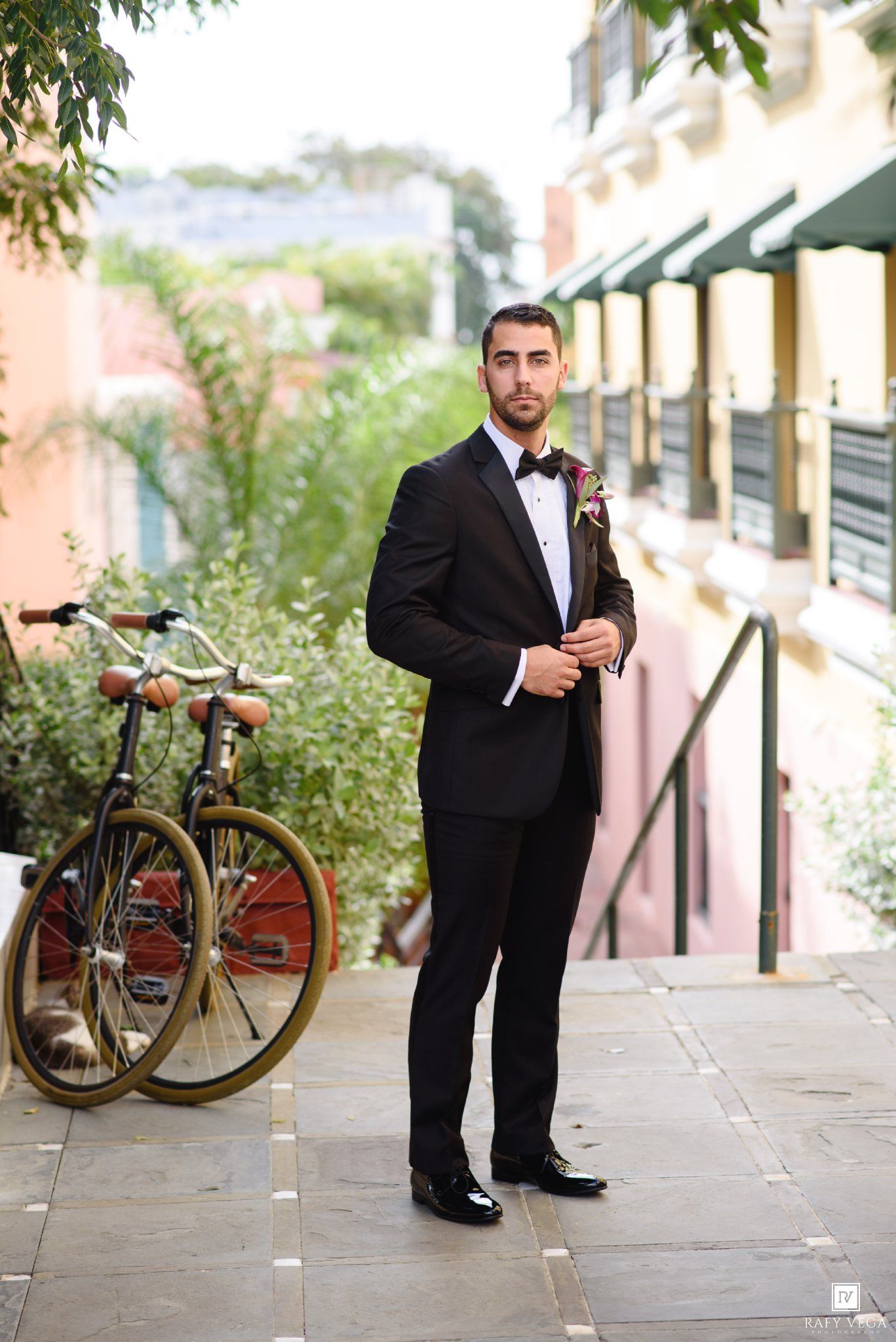 Groom in black tuxedo with bow tie poses outdoors near vintage bicycle and colorful buildings.