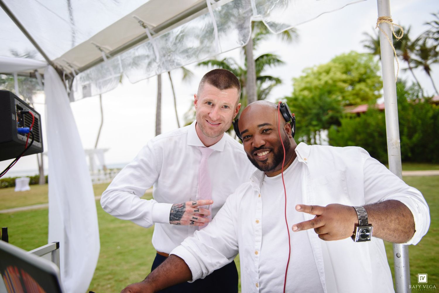 Two men in white shirts smile together at an outdoor DJ setup with palm trees in the background.
