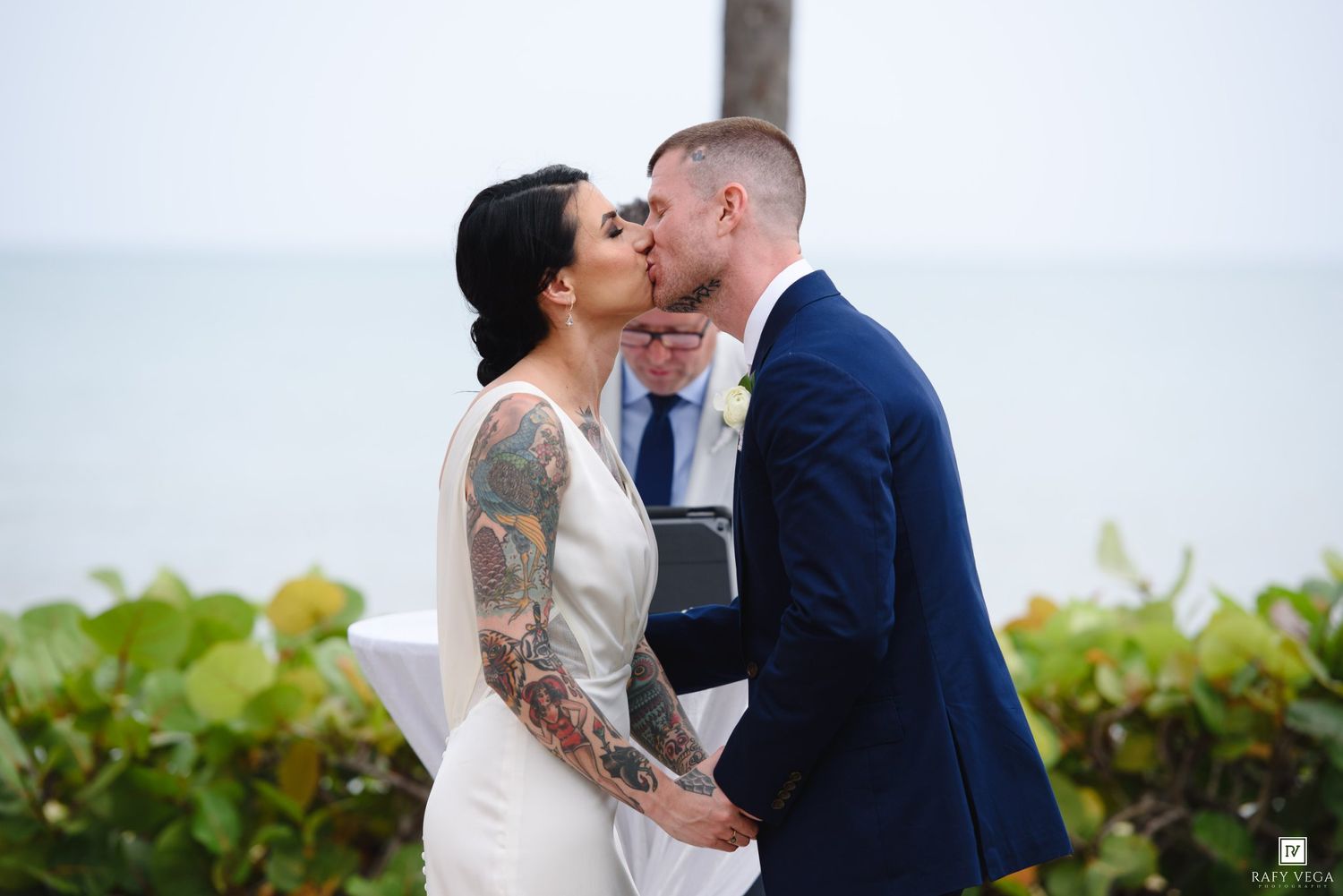Couple sharing first kiss at outdoor beach wedding ceremony, bride with tattoos in white gown, groom in navy suit.